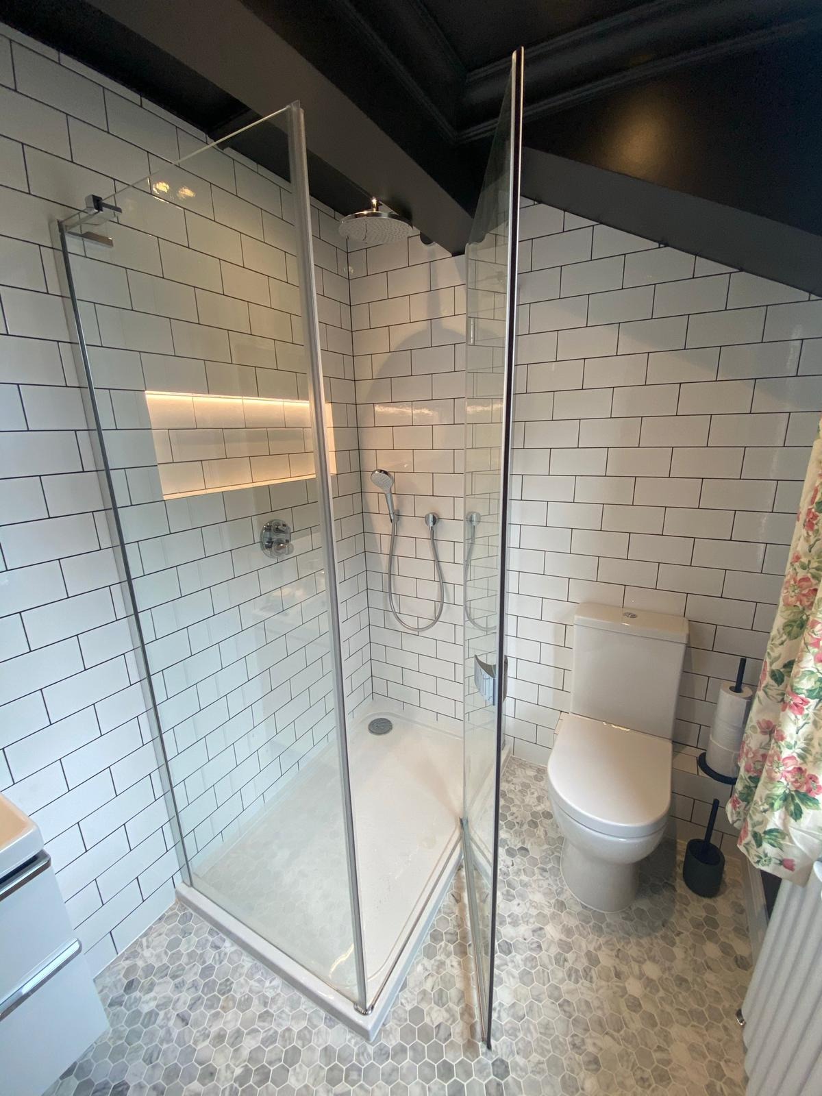 Nowoczesna łazienka z kabiną prysznicową typu walk-in, białymi płytkami imitującymi cegłę, szarą mozaiką na podłodze i podświetlaną wnęką w ścianie.