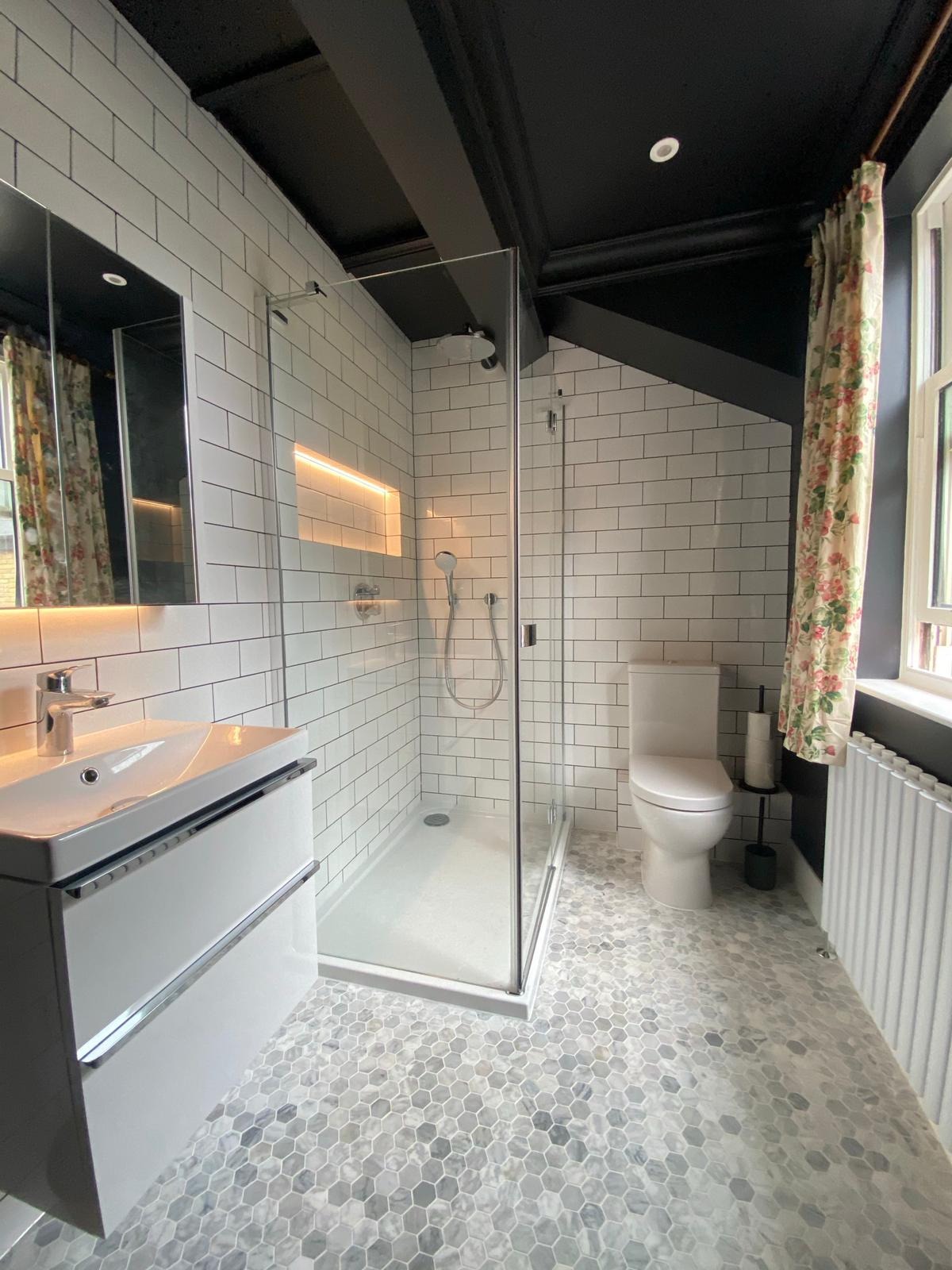 Nowoczesna łazienka z białą armaturą, kabiną prysznicową ze szklanymi drzwiami, umywalką z szafką, heksagonalną mozaiką na podłodze i akcentami w postaci czarnego sufitu z belkami oraz zasłony...