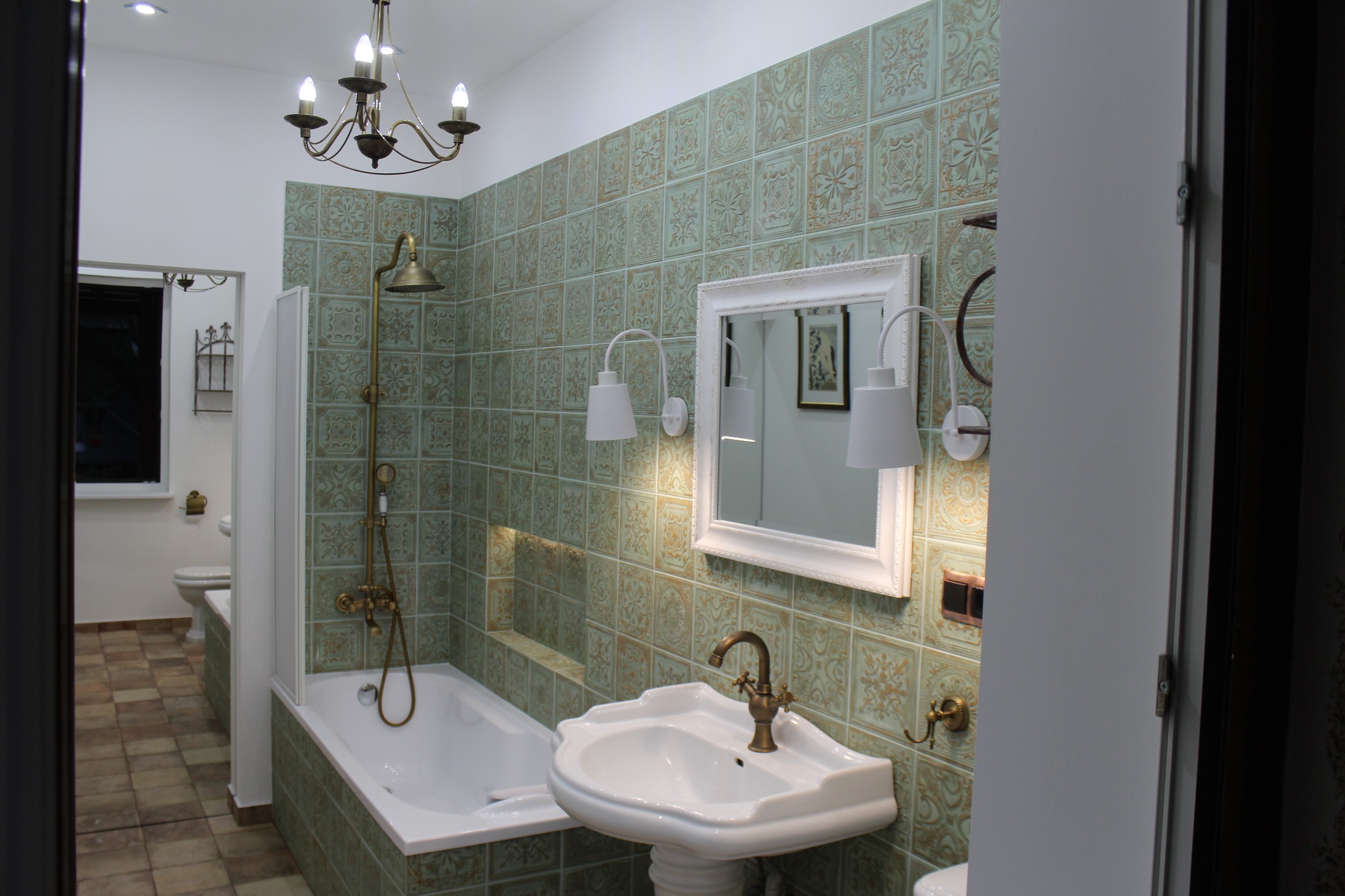 Retro łazienka z zielono-złotymi, wzorzystymi płytkami, białą armaturą i baterią w stylu vintage, oświetlona kinkietami i żyrandolem.