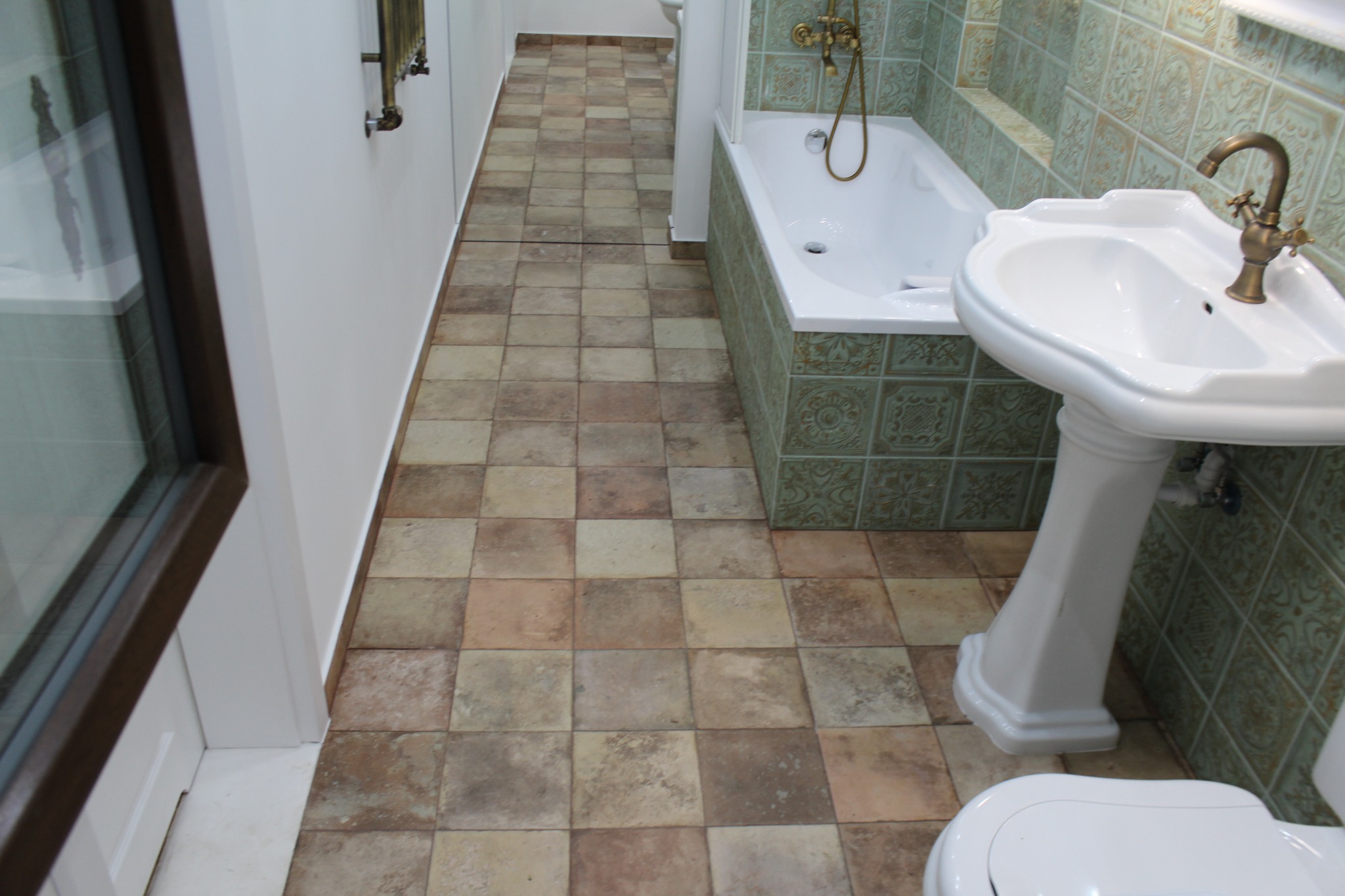 Umywalka na postumencie z baterią w stylu retro, wanna obudowana płytkami z dekoracyjnym wzorem oraz podłoga wyłożona kwadratowymi płytkami w odcieniach beżu i brązu w łazience.