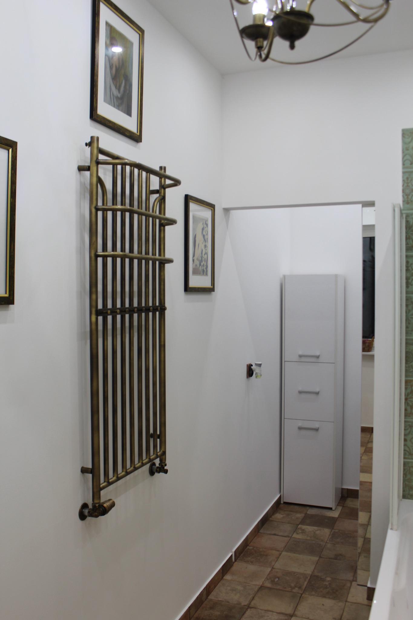 Korytarz z białymi ścianami, terakotową podłogą i grzejnikiem drabinkowym w stylu retro, ozdobiony obrazami w złotych ramach i oświetlony ozdobnym żyrandolem.