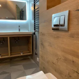 Nowoczesna łazienka z drewnopodobną okładziną ścienną, chromowanym przyciskiem spłuczki, podświetlanym lustrem i grzejnikiem drabinkowym, widoczna również częściowo umywalka i toaleta.