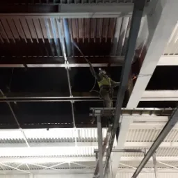 Pracownik w kombinezonie ochronnym maluje natryskowo sufit hali przemysłowej z metalową konstrukcją, stojąc na rusztowaniu.