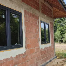 Dwa okna z czarnymi ramami osadzone w surowej ścianie z czerwonej cegły, widoczne ślady pianki montażowej i świeżego tynku, tło stanowią drzewa i błękitne niebo.