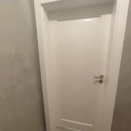 Zamknięte, białe drzwi wewnętrzne z ramą, klamka w kolorze złotym, szara ściana w tle.