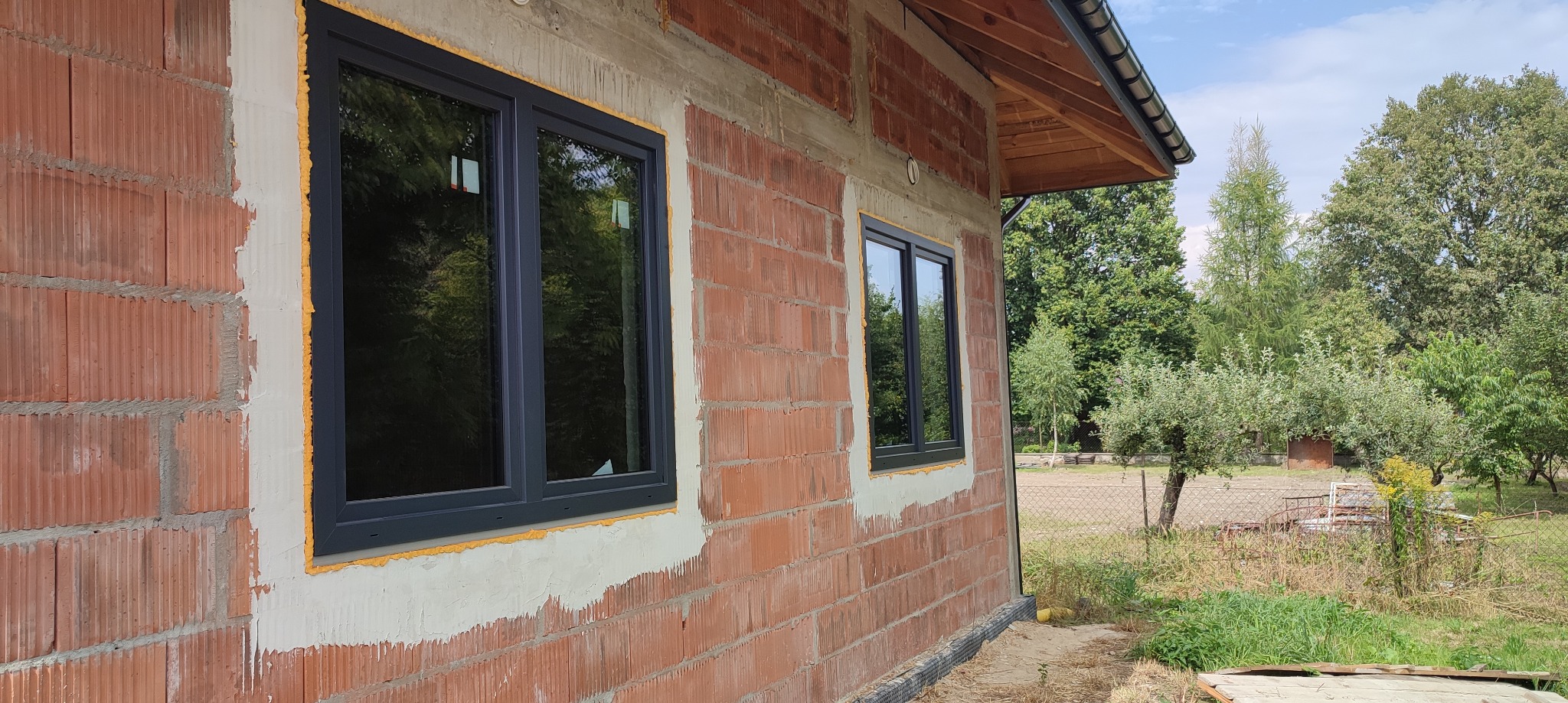 Dwa okna z czarnymi ramami osadzone w surowej ścianie z czerwonej cegły, widoczne ślady pianki montażowej i świeżego tynku, tło stanowią drzewa i błękitne niebo.