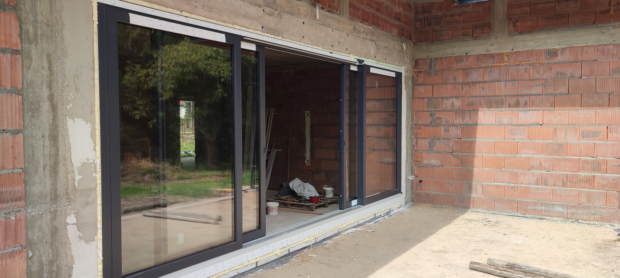 Montaż przesuwnych drzwi balkonowych z ciemną ramą w surowym stanie budynku, widoczne cegły i materiały budowlane wewnątrz i na zewnątrz.