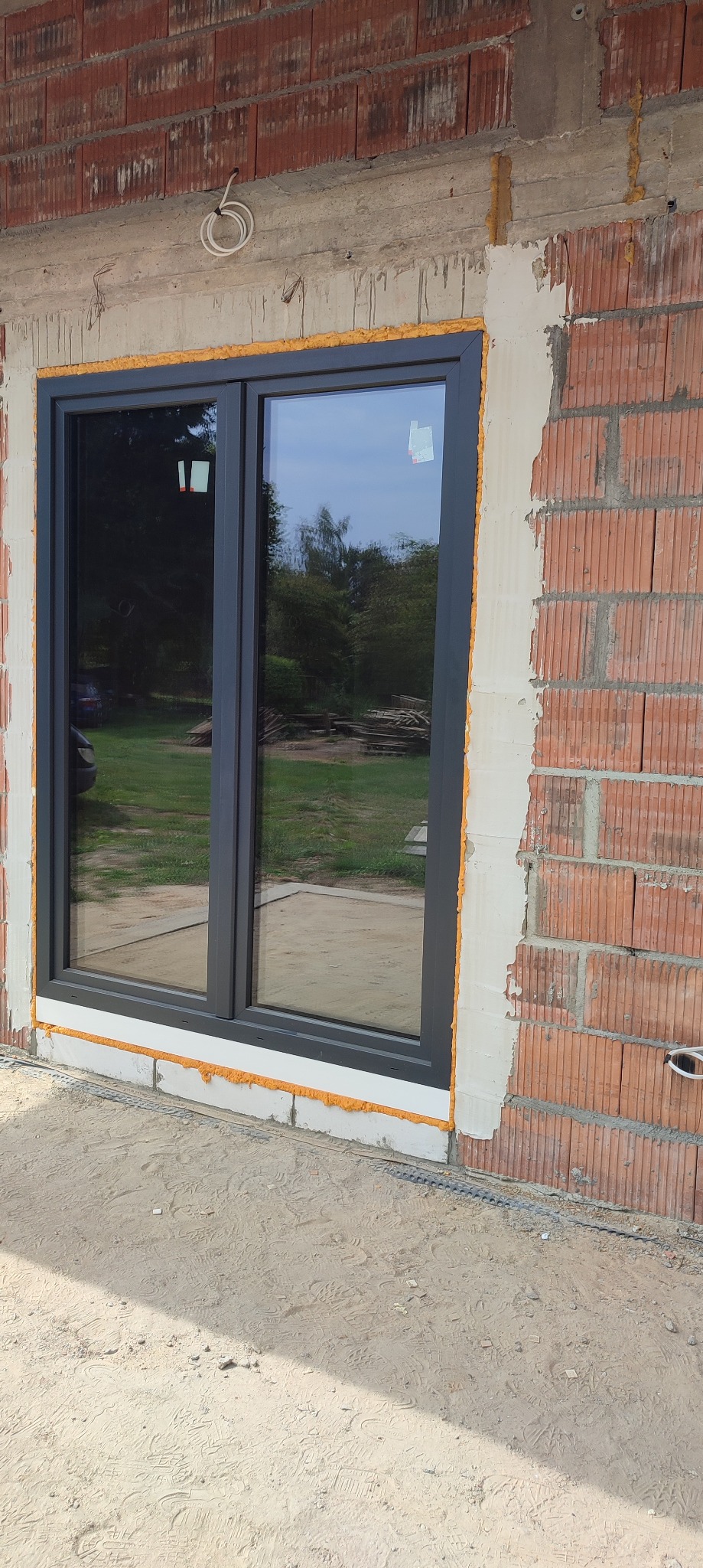 Szare dwuskrzydłowe okno w trakcie montażu w surowej ścianie z czerwonej cegły, widoczna pianka montażowa i częściowo nałożony tynk.