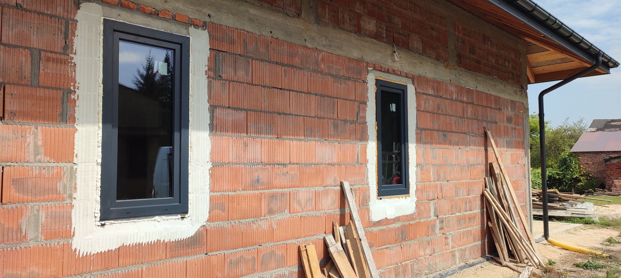 Dwa okna z grafitowymi ramami osadzone w murze z czerwonej cegły, widoczne ślady pianki montażowej i zaprawy, w tle fragment dachu i rynny.