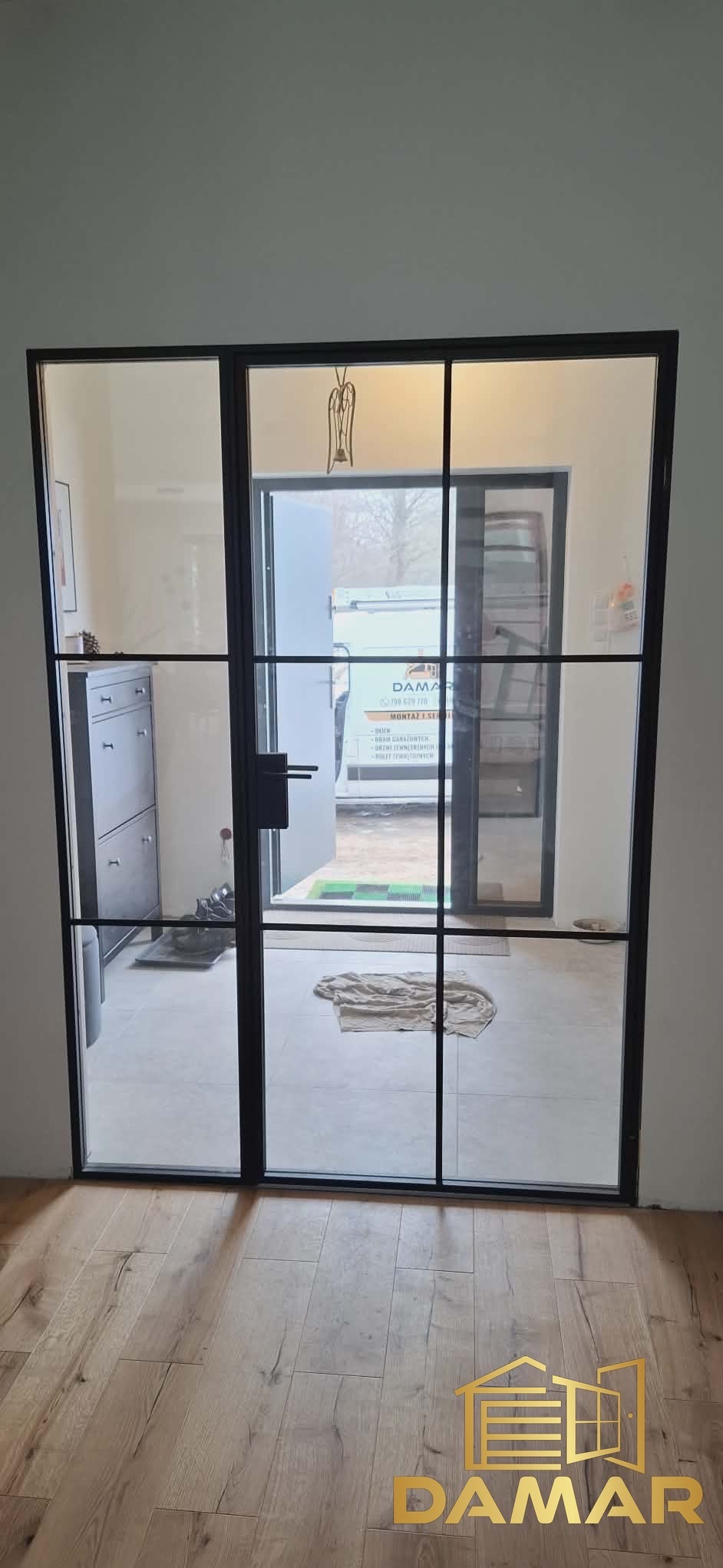 Nowoczesne, czarne drzwi zewnętrzne z przeszkleniami w stylu loftowym. Widoczny fragment wnętrza i podjazd z samochodem dostawczym.