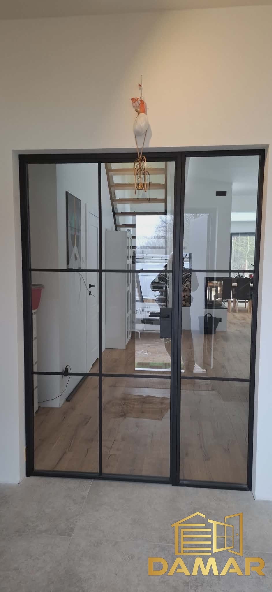 Nowoczesne, przesuwne drzwi wewnętrzne z czarnymi szprosami, oddzielające przestrzeń z widocznymi schodami i salonem. Minimalistyczny design, jasne wnętrze.