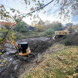 DEJV-BUD Dawid Trzebniak - Dwa żółte, gąsienicowe mini-koparki CAT pracują w błotnistym wykopie, jesienne liście na pierwszym planie, drzewa i budynki w tle, pochmurne niebo.