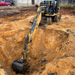 DEJV-BUD Dawid Trzebniak - Żółta koparka CAT w trakcie wykopu głębokiego, okrągłego dołu w ziemi. Operator w kabinie. W tle budynek z czerwoną dachówką i ogrodzenie.