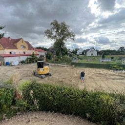 DEJV-BUD Dawid Trzebniak - Żółta mini koparka CAT na wyrównanym terenie budowy, widok z góry na tle domów i pochmurnego nieba. Prace ziemne przygotowawcze pod budowę.