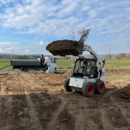 Biała miniładowarka Bobcat S175 z pełną łyżką ziemi na placu budowy, w tle wywrotka Mercedes-Benz z HDS oraz zielone pola i błękitne niebo z chmurami.