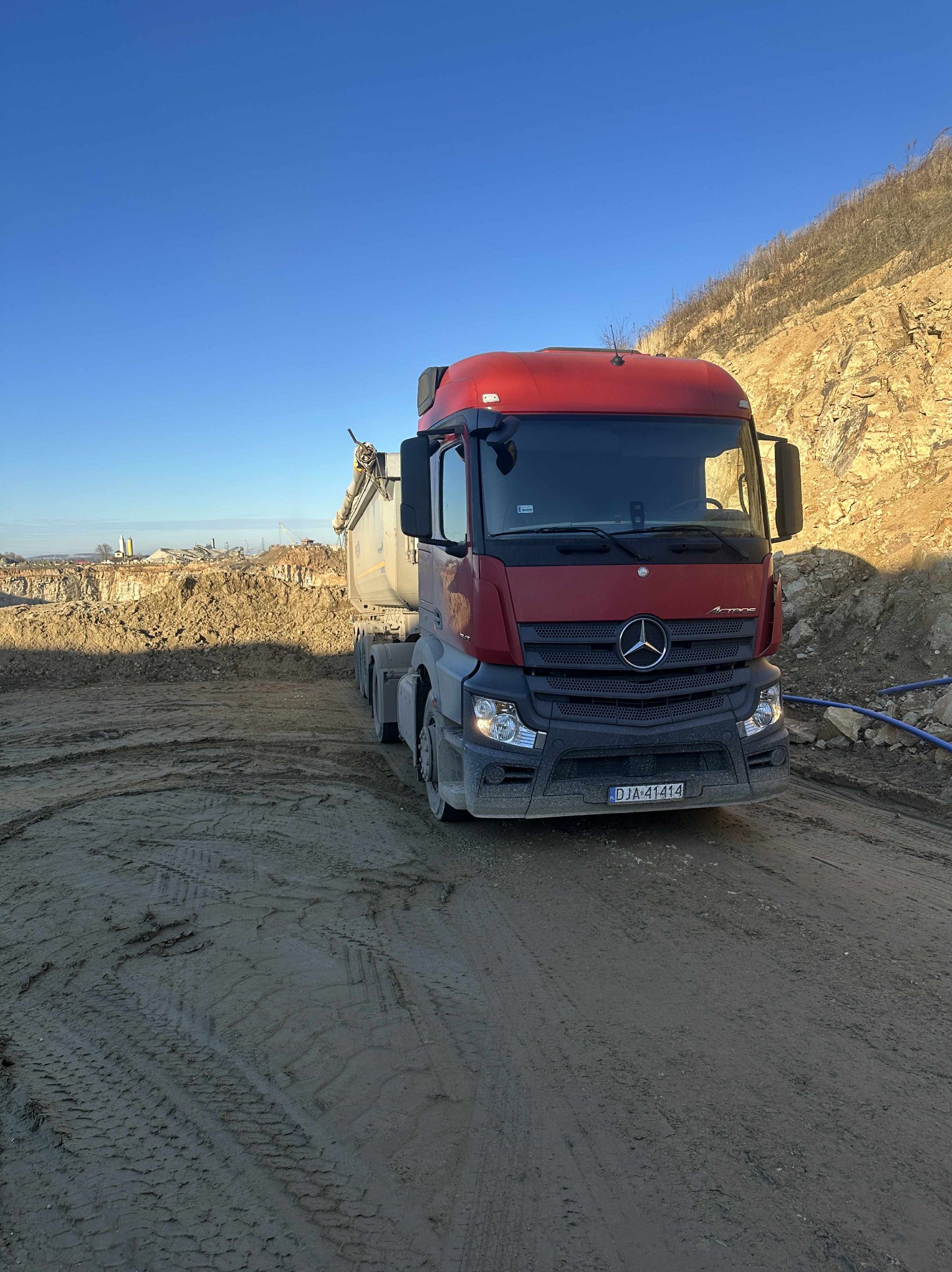 Czerwona ciężarówka Mercedes na placu budowy w Drzymałowicach, z wykopem i stertą ziemi w tle, pod błękitnym niebem. Widoczne ślady opon na błotnistym podłożu.