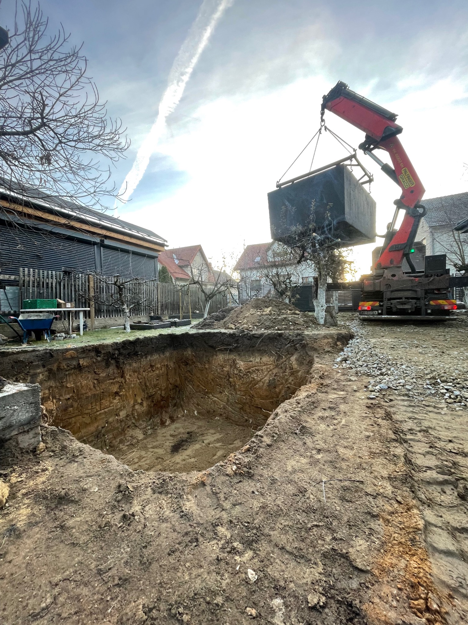 Montaż betonowego szamba w wykopie za pomocą dźwigu samochodowego na tle zabudowy jednorodzinnej i drzew bez liści.