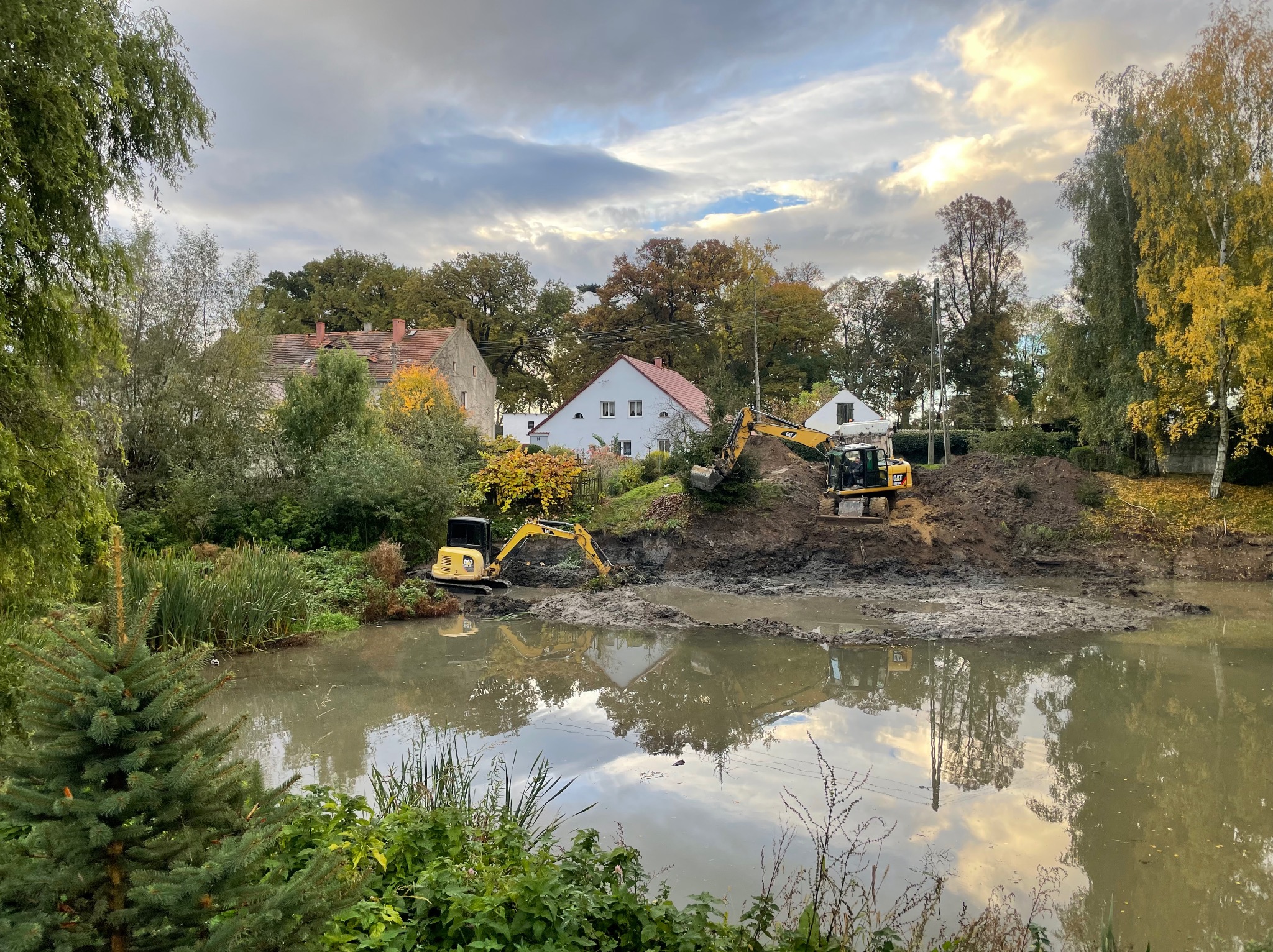 Dwie żółte koparki pracują na brzegu mulistego stawu, w tle domy i drzewa pod pochmurnym niebem. Prace ziemne w toku, odbicie w wodzie.