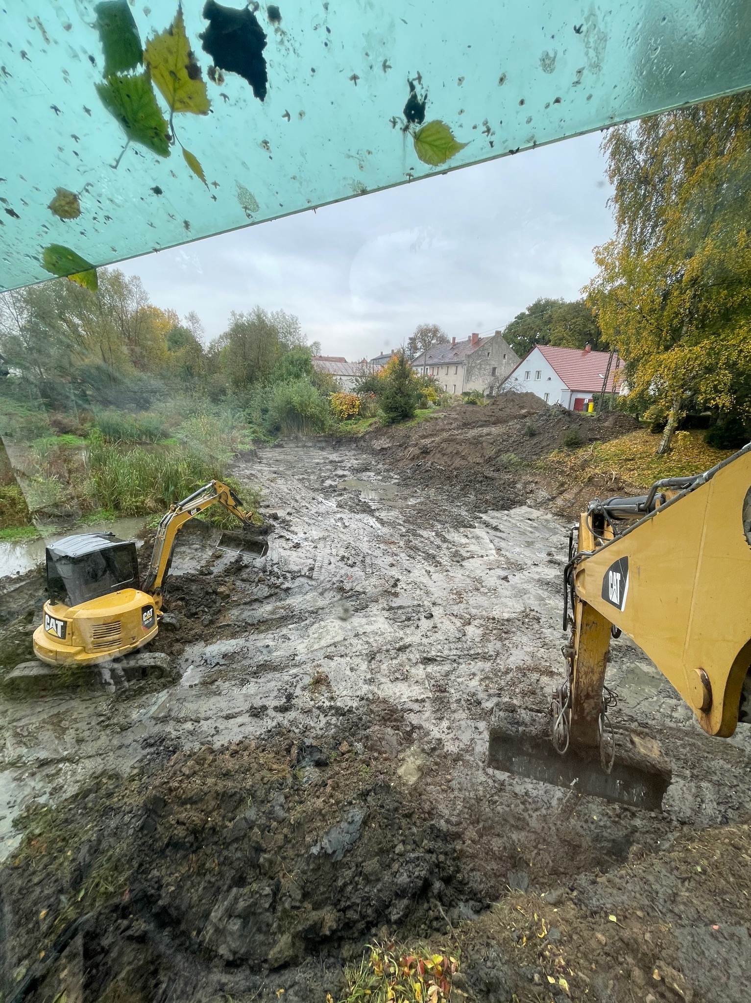 Dwa żółte mini-koparki CAT pracują na błotnistym terenie, przygotowując teren pod budowę. Widok zza szyby z liśćmi, w tle zabudowania.