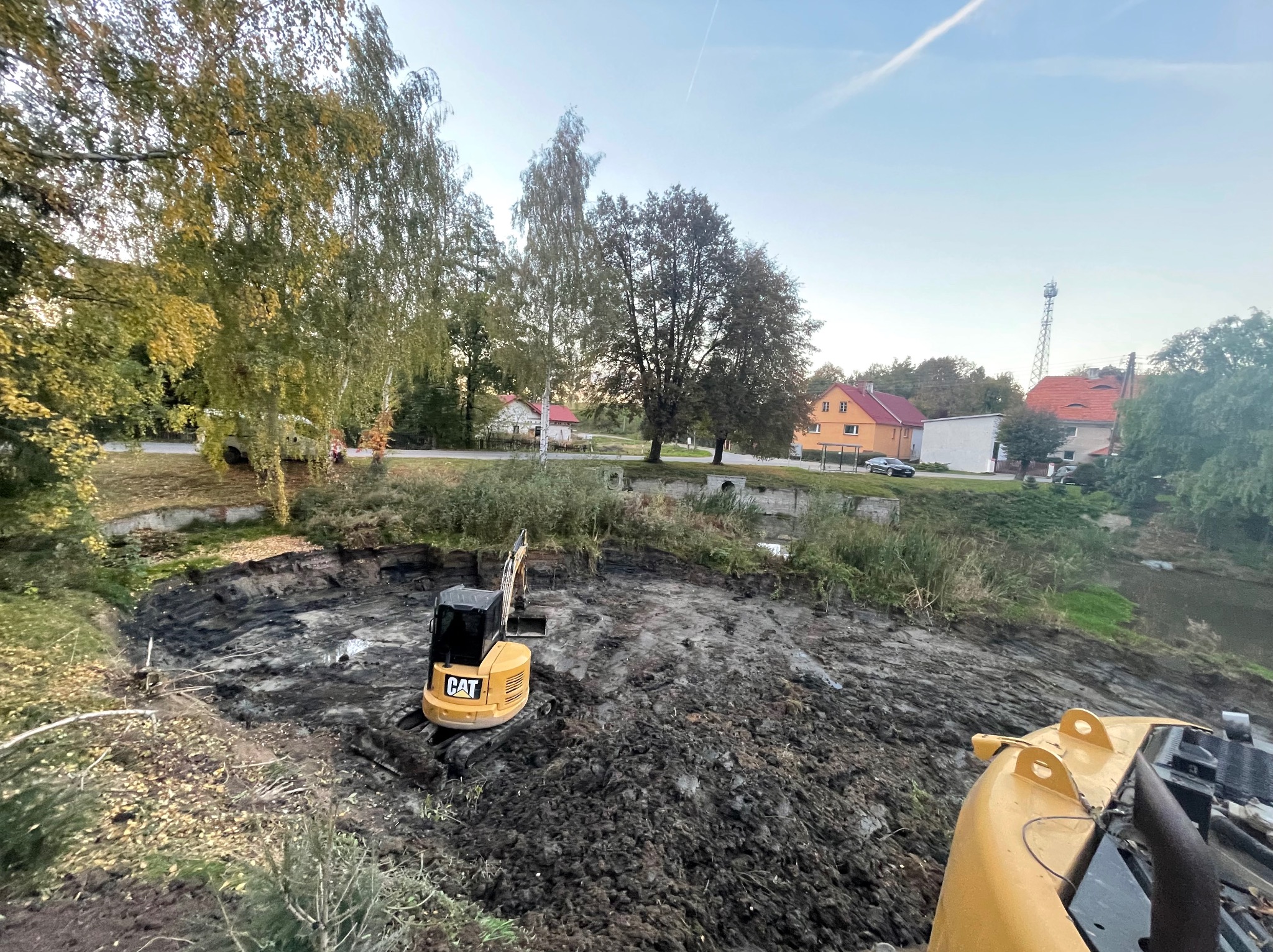 Żółta koparka CAT w wykopie ziemnym, prawdopodobnie przygotowanie terenu pod budowę. W tle drzewa, budynki mieszkalne i wieża telekomunikacyjna.