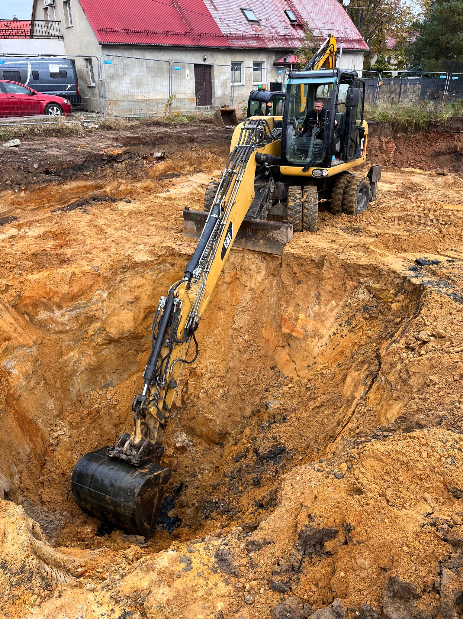 Żółta koparka CAT w trakcie wykopu głębokiego, okrągłego dołu w ziemi. Operator w kabinie. W tle budynek z czerwoną dachówką i ogrodzenie.
