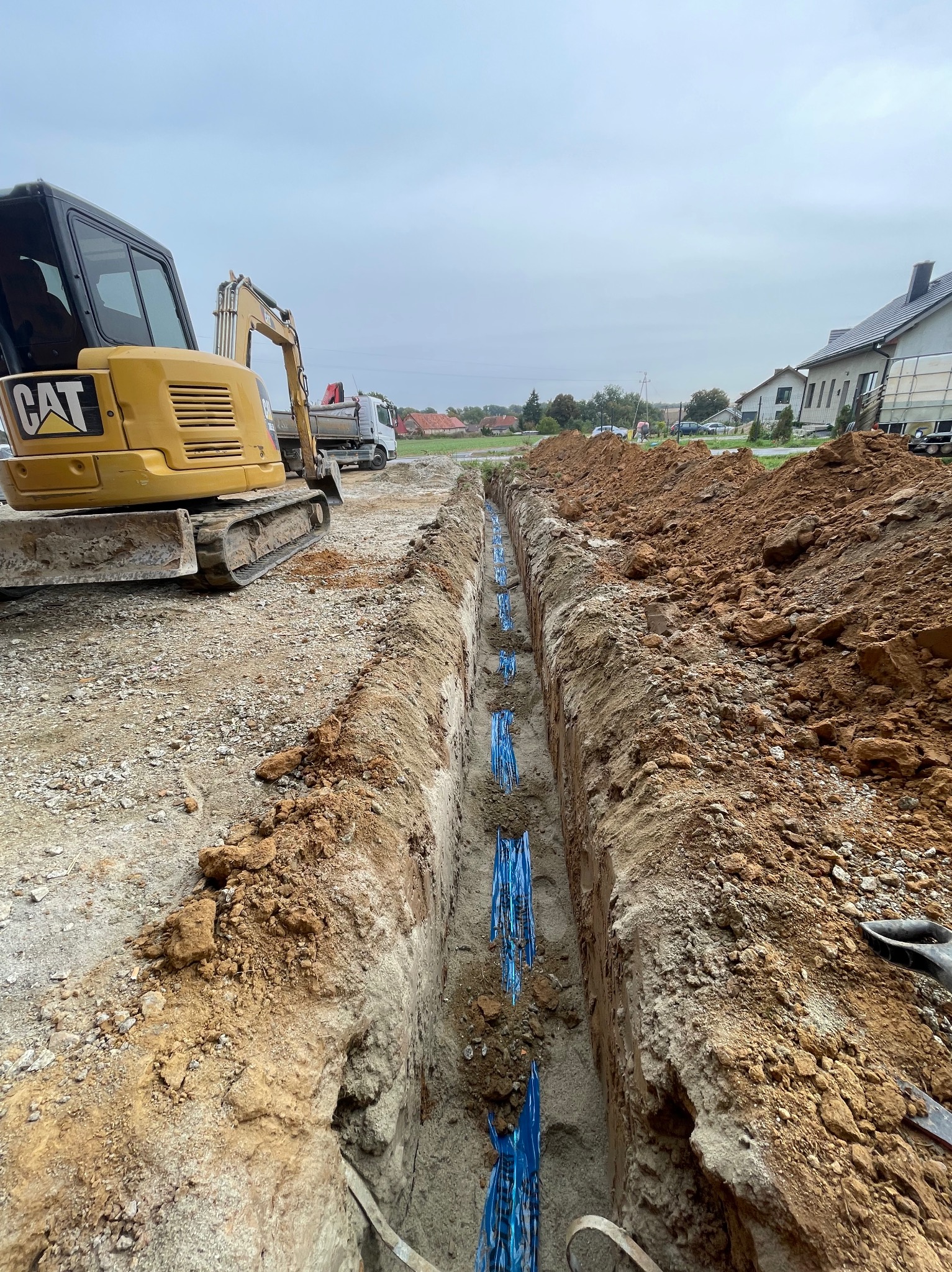 Wykopany rów z niebieskimi rurami w ziemi, obok żółta koparka CAT i hałda ziemi. Widok na budynki w tle. Prace ziemne, instalacje podziemne.