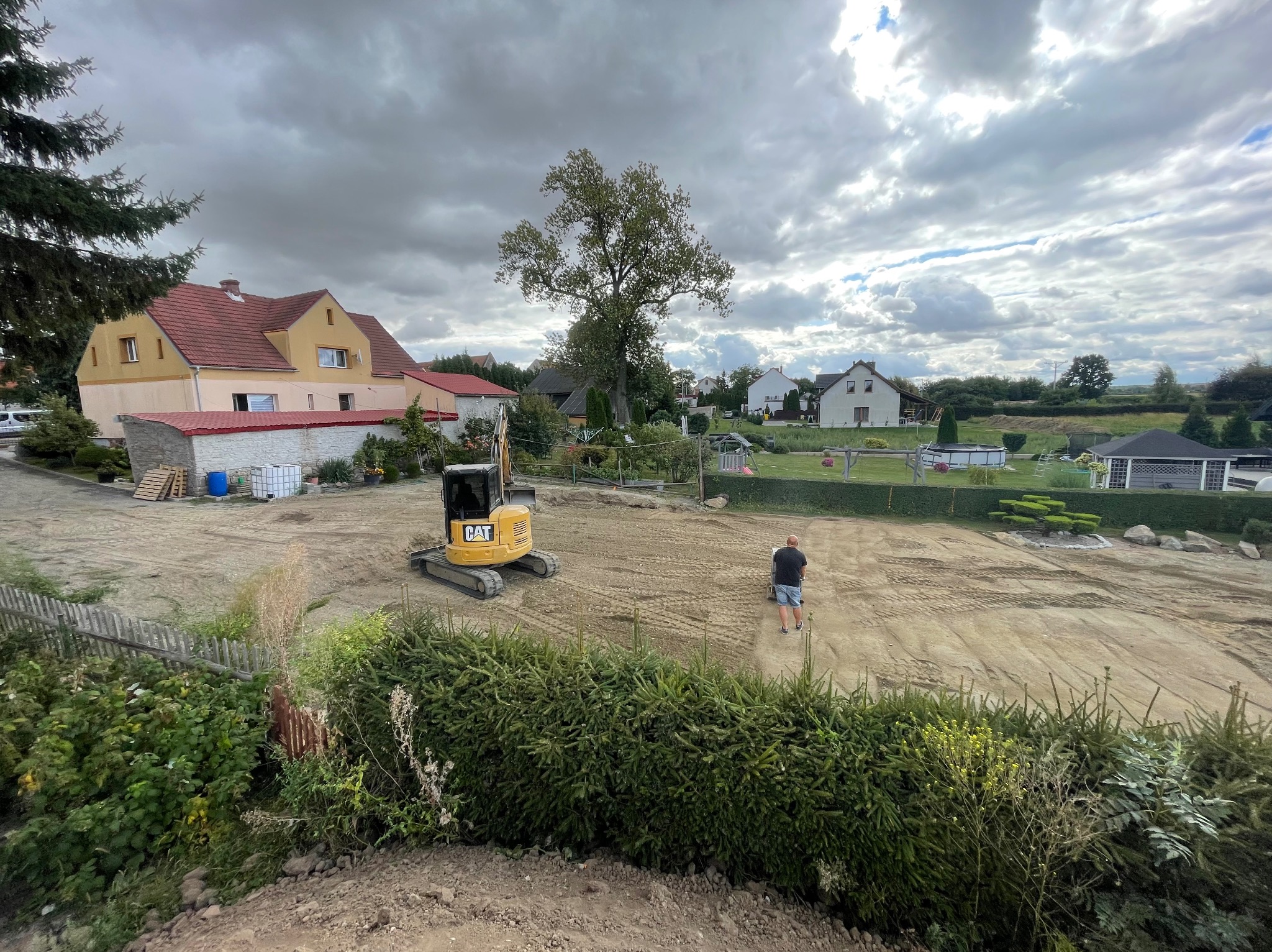 Żółta mini koparka CAT na wyrównanym terenie budowy, widok z góry na tle domów i pochmurnego nieba. Prace ziemne przygotowawcze pod budowę.