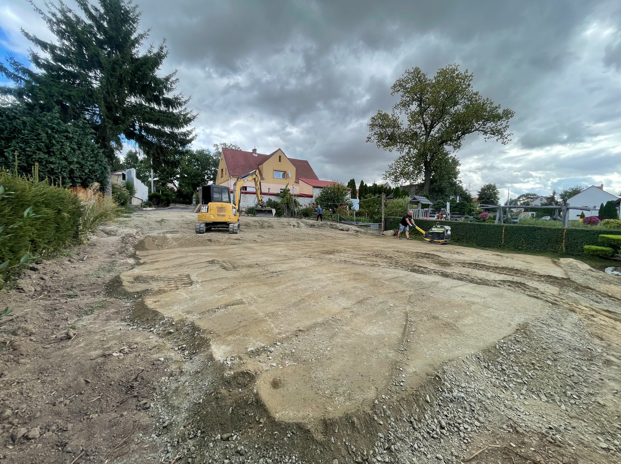 Żółta minikoparka i zagęszczarka na wyrównanym terenie pod budowę domu jednorodzinnego, widok na budynek i pochmurne niebo.