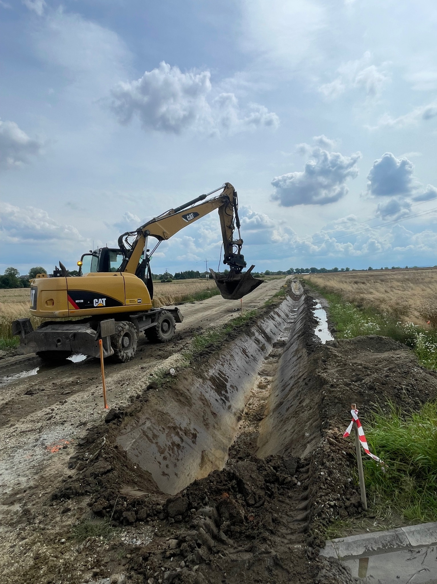 Żółta koparka CAT M313D pogłębia rów melioracyjny na polu, widok z boku, w tle niebo z chmurami. Rów wypełniony wodą, w oddali widoczne drzewa.