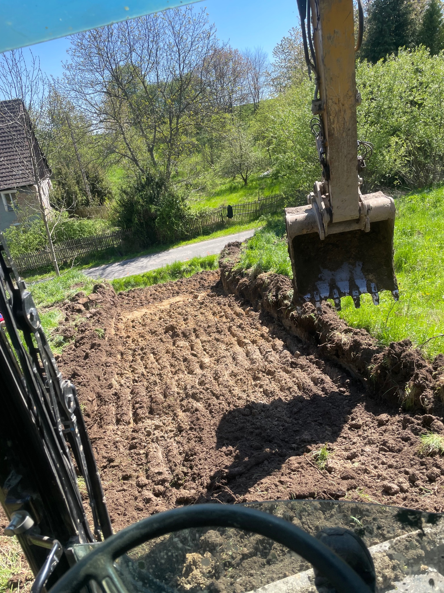 Żółta koparka w trakcie wykopu wąskiego rowu w ziemi na zielonym terenie, widok z kabiny operatora, w tle dom i drzewa.