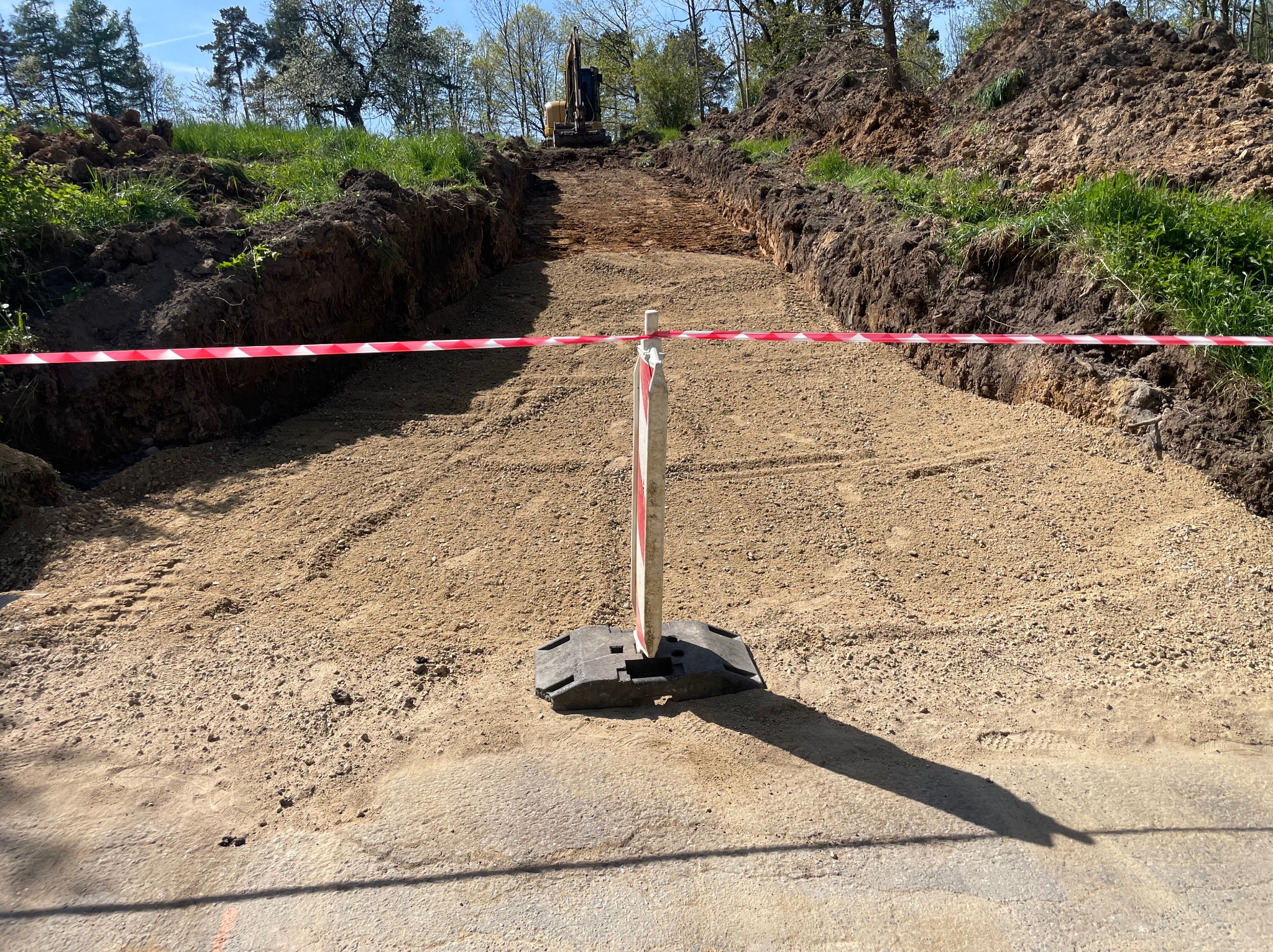Wykop pod drogę z wykopaną ziemią po bokach, zabezpieczony taśmą ostrzegawczą i znakiem. W tle widoczna koparka i drzewa.