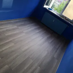 Pokój po remoncie: świeżo pomalowane na niebiesko ściany, szare panele podłogowe ułożone w jodełkę, niebieski grzejnik pod oknem z widokiem na drzewa i budynki w oddali.