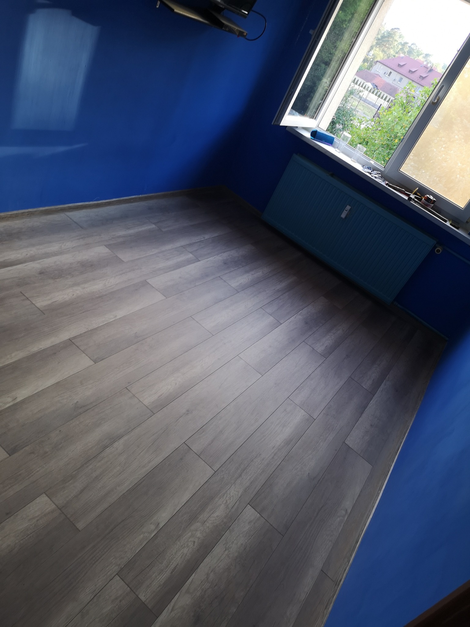 Pokój po remoncie: świeżo pomalowane na niebiesko ściany, szare panele podłogowe ułożone w jodełkę, niebieski grzejnik pod oknem z widokiem na drzewa i budynki w oddali.