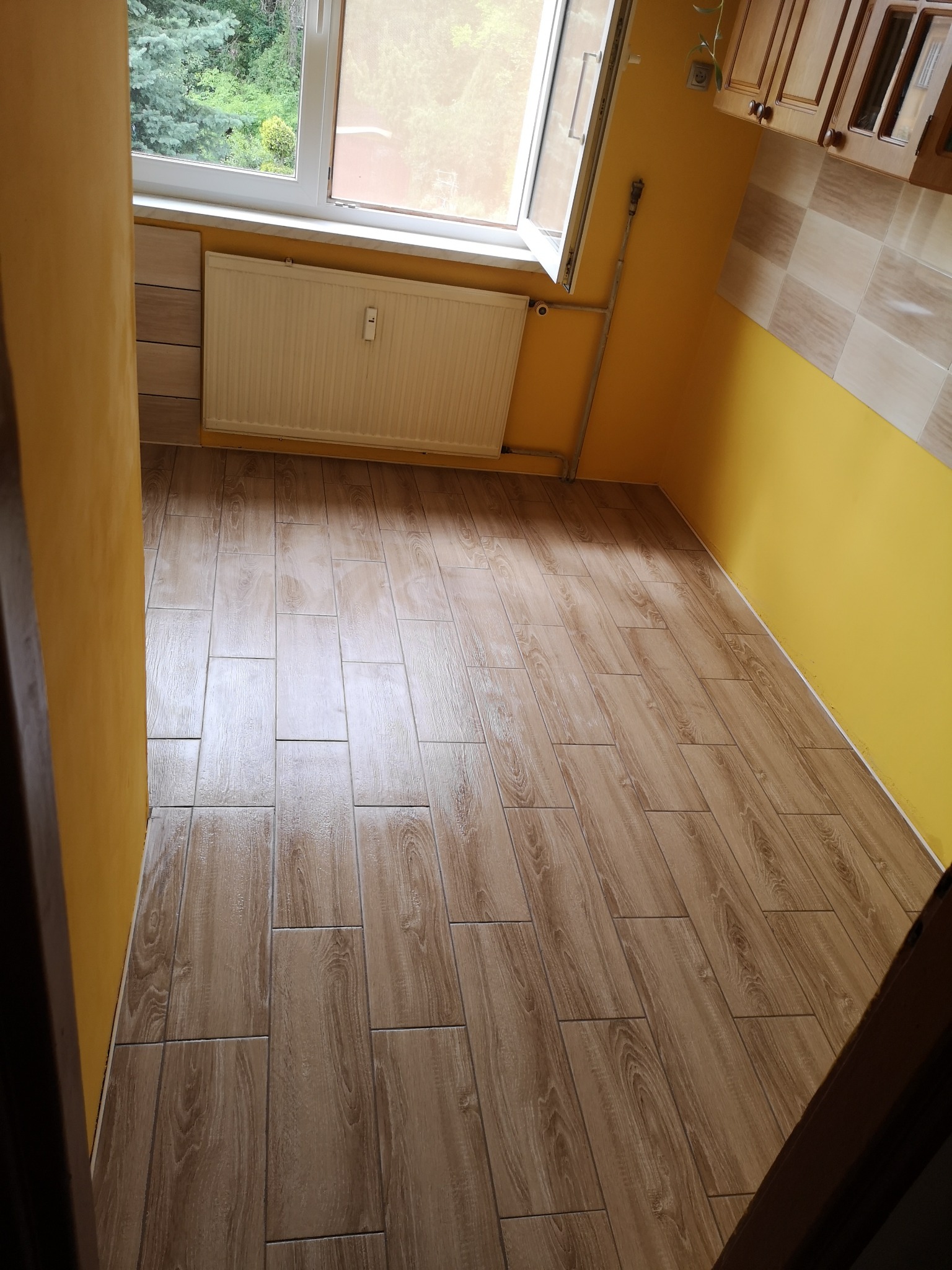 Wykończona podłoga z płytek imitujących drewno w małej kuchni z żółtymi ścianami i białym grzejnikiem pod oknem.