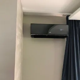 Czarny klimatyzator AUX zamontowany na ścianie w jasnym pomieszczeniu, obok zasłoniętego okna z granatowymi zasłonami.