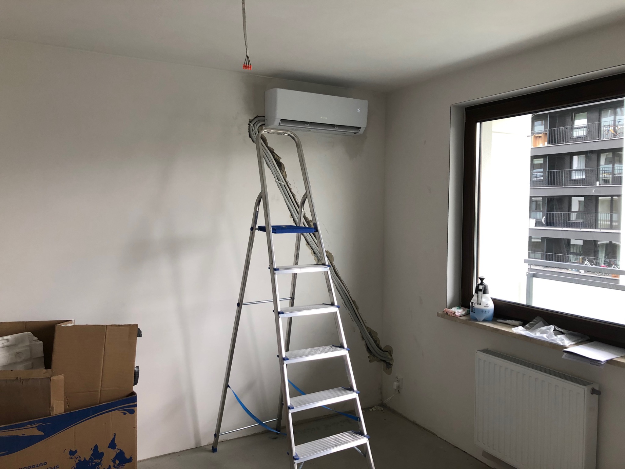 Nowoczesny klimatyzator zamontowany na ścianie w jasnym pomieszczeniu, widoczna drabina aluminiowa, odsłonięte przewody, kartony na podłodze, okno z widokiem na blok mieszkalny.