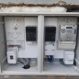 Otwarta skrzynka elektryczna z zainstalowanym licznikiem, bezpiecznikami i przewodami, widoczne elementy montażowe i fragment ściany zewnętrznej.