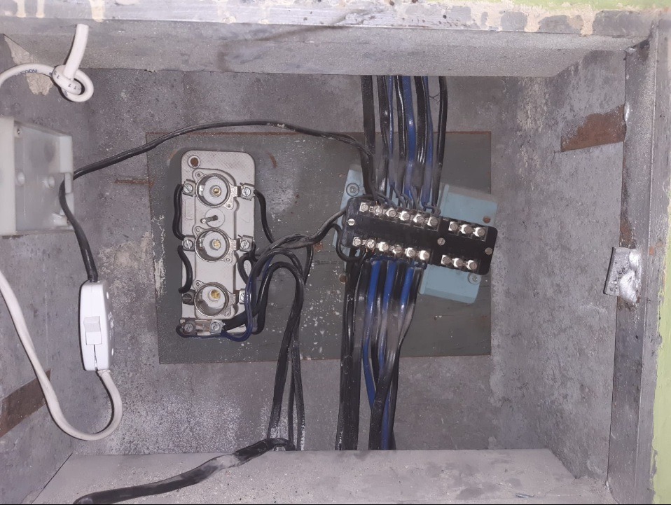 Wnętrze betonowej skrzynki z zamontowanymi elementami instalacji elektrycznej: gniazdo, przełącznik, listwa zaciskowa z niebieskimi przewodami i ceramiczna kostka. Widoczne ślady kurzu...