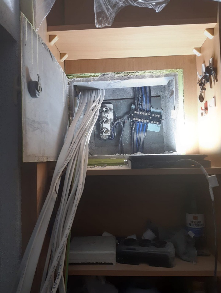Otwarta szafka z widoczną instalacją elektryczną: liczne białe kable prowadzące do skrzynki z bezpiecznikami i złączkami, tło stanowią półki z różnymi przedmiotami.