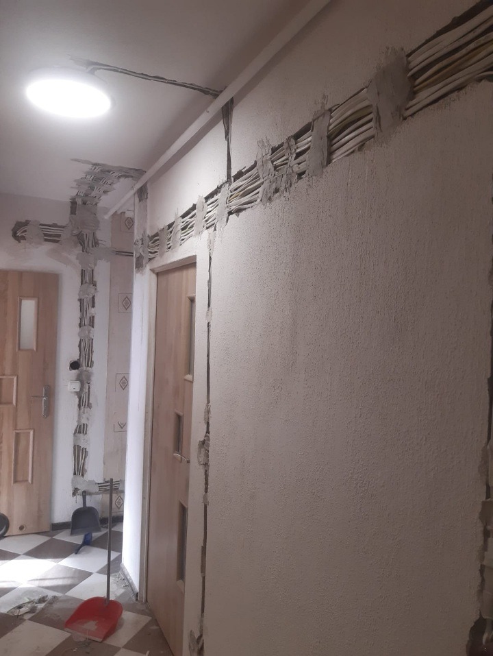 Wnętrze w trakcie remontu z widocznymi przewodami elektrycznymi osadzonymi w ścianie i suficie, częściowo zamaskowanymi gipsem, z widoczną podłogą w szachownicę i drzwiami w tle.