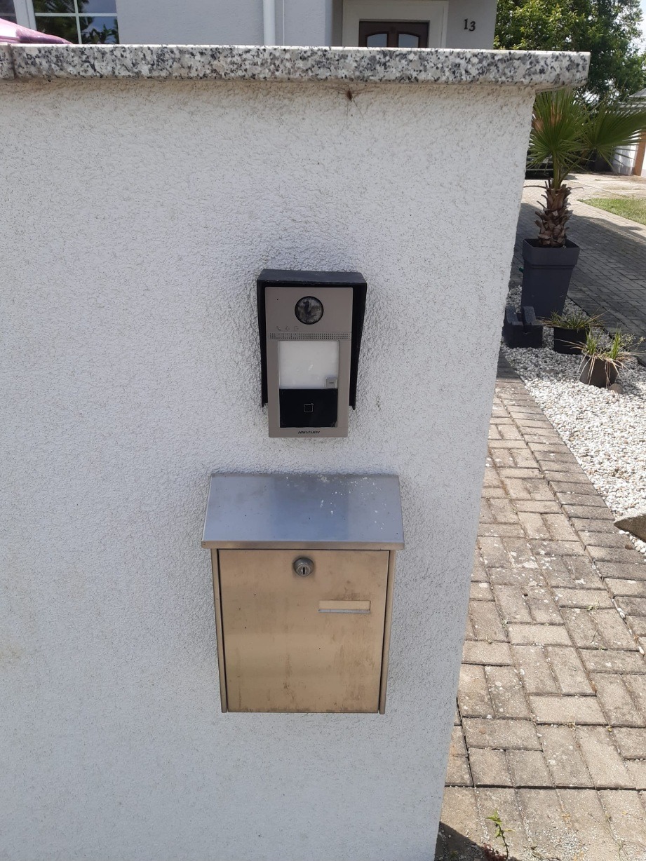 Domofon z kamerą i metalowa skrzynka pocztowa zamontowane na białym, teksturowanym murze przy ścieżce z kostki brukowej.