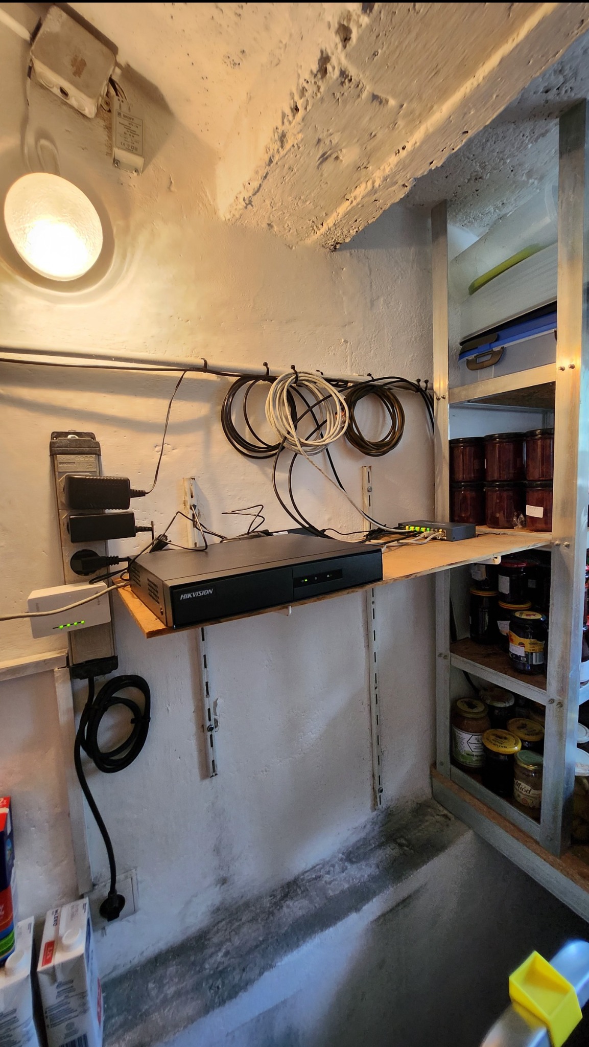 Zamontowany rejestrator monitoringu Hikvision z podłączonymi kablami sieciowymi i zasilaczami, umieszczony na drewnianej półce w piwnicy obok metalowego regału z przetworami.