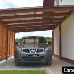 Szary samochód Nissan Qashqai zaparkowany pod zadaszeniem z brązowego drewna i przezroczystego poliwęglanu, z boczną ścianą z żaluzji w kolorze pomarańczowym.