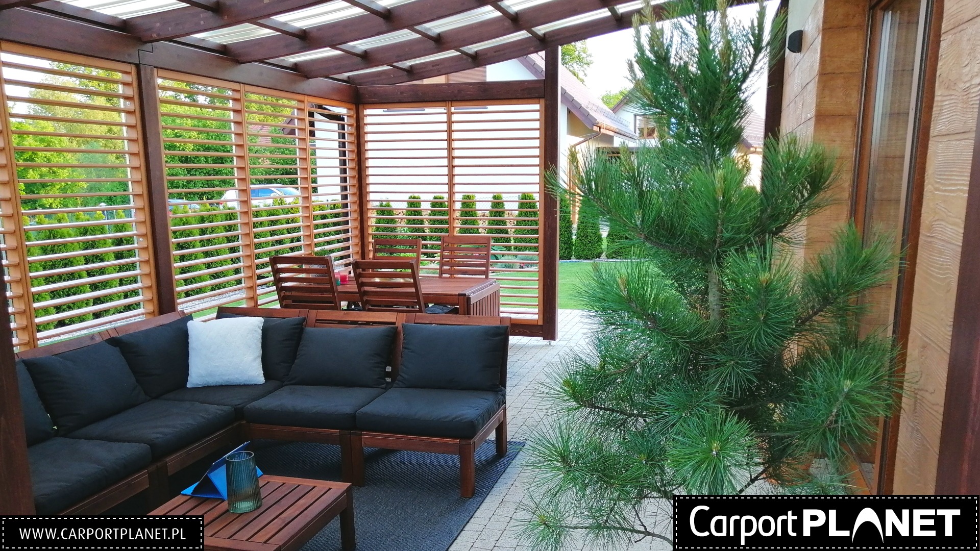 Drewniana pergola z lamelami, wyposażona w ciemne meble wypoczynkowe i stół z krzesłami, widok z zewnątrz przesłonięty iglakiem, w tle zadbany ogród.