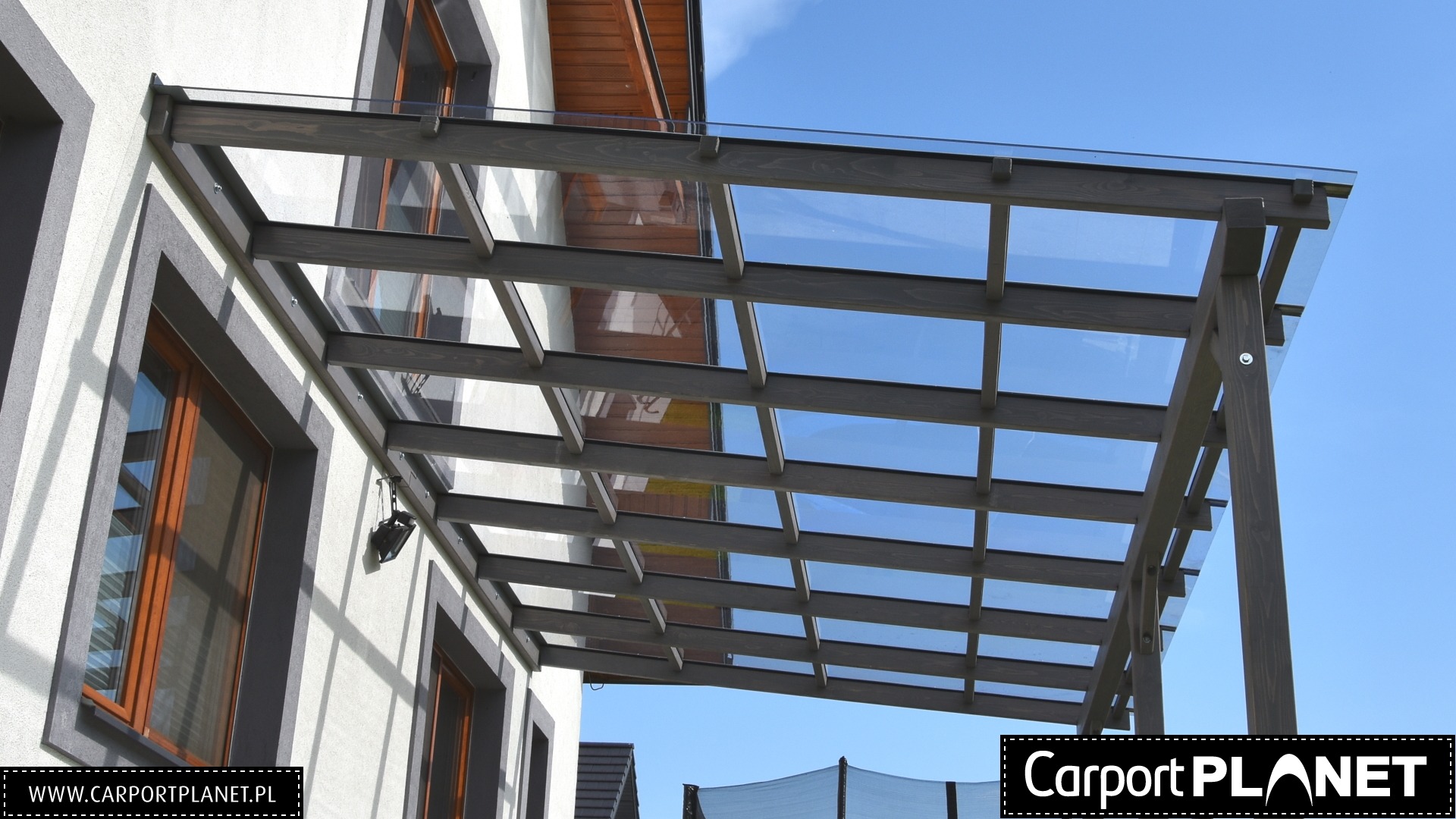 Nowoczesna pergola przyścienna z drewnianą konstrukcją i szklanym dachem, widoczna z dołu pod kątem, z logo firmy w prawym dolnym rogu.