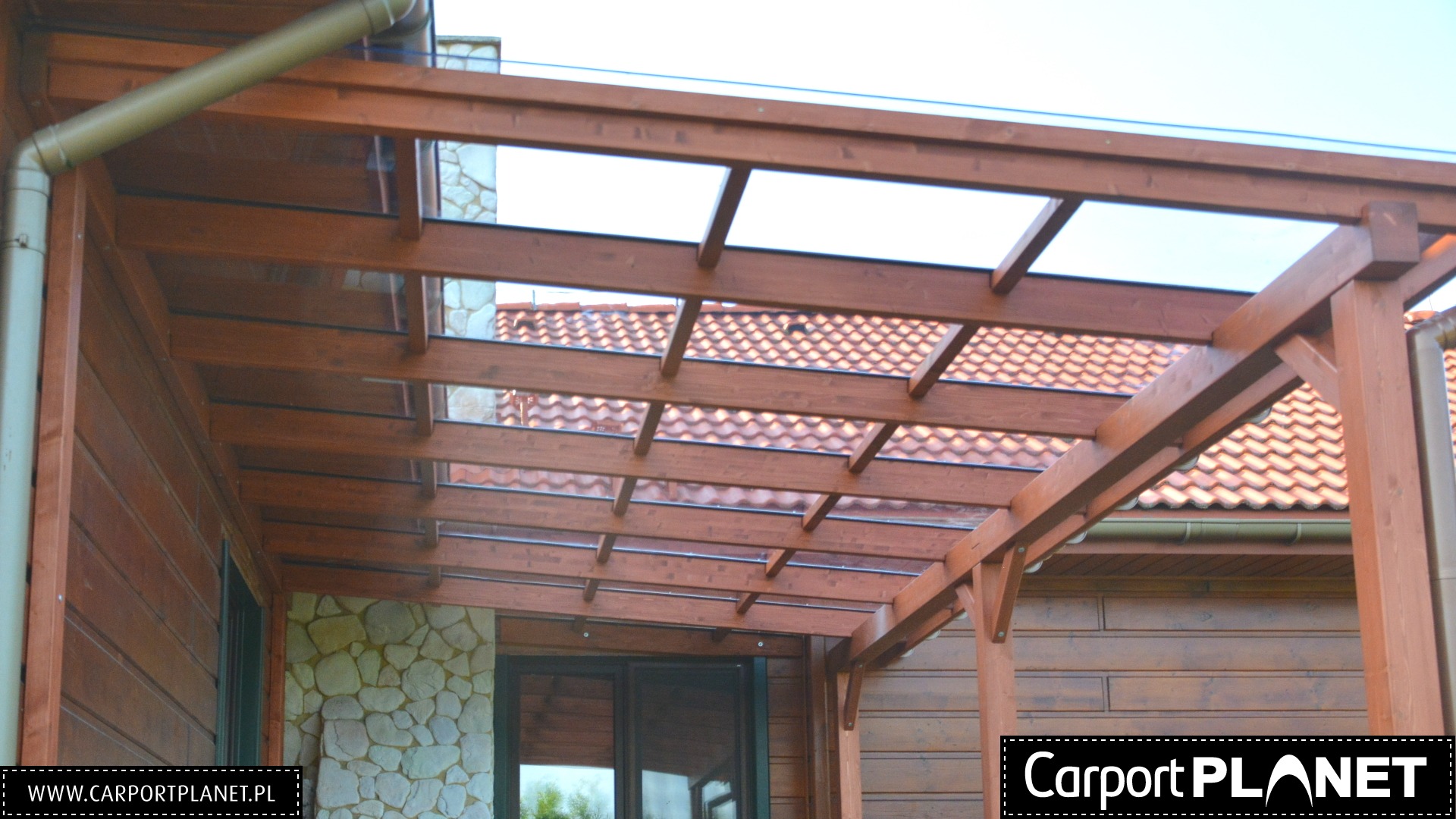 Drewniana pergola z przezroczystym zadaszeniem przylegająca do domu z elewacją z kamienia i drewnianych paneli.