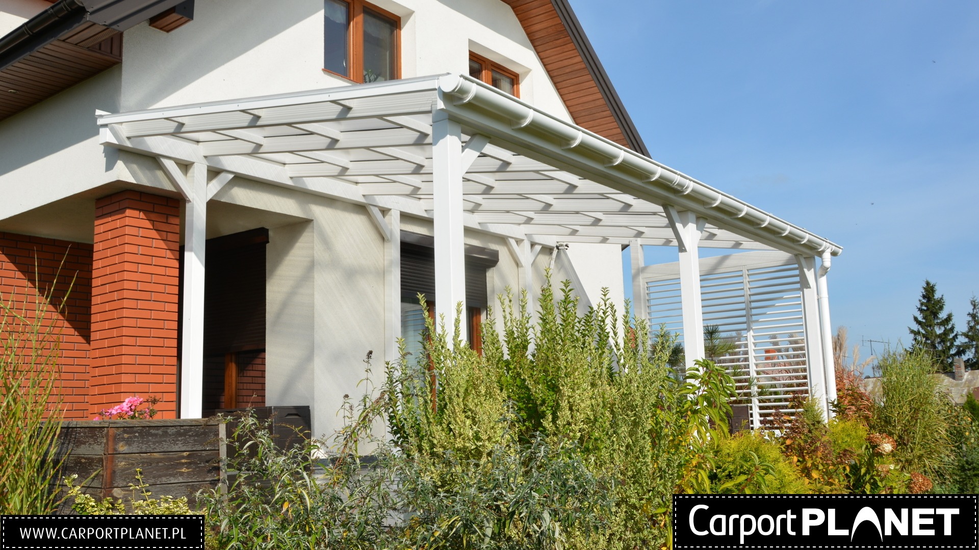 Biała konstrukcja z zadaszeniem przylegająca do domu z ceglaną kolumną, widoczne rośliny w ogrodzie.