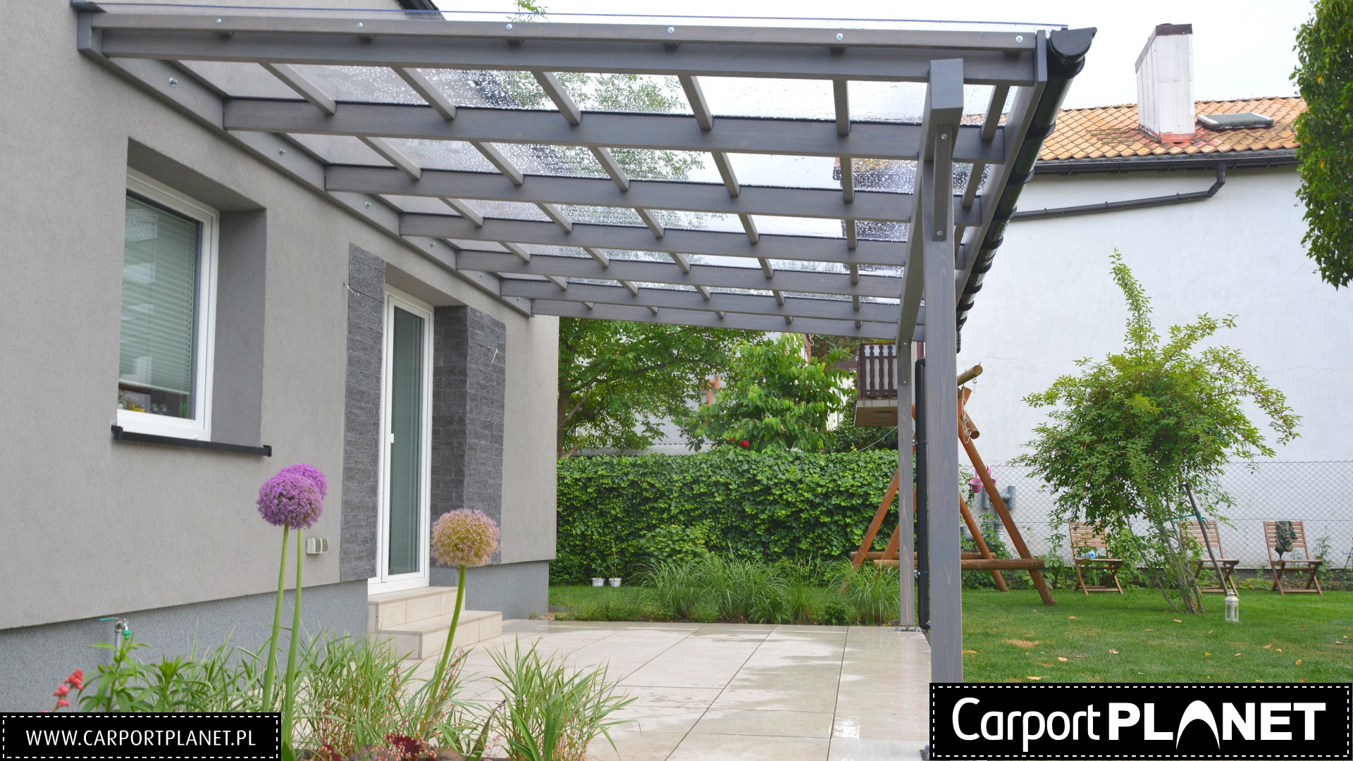 Szara konstrukcja zadaszenia tarasu z przezroczystym dachem, widok na ogród z huśtawką i krzesłami, fioletowe kwiaty na pierwszym planie.