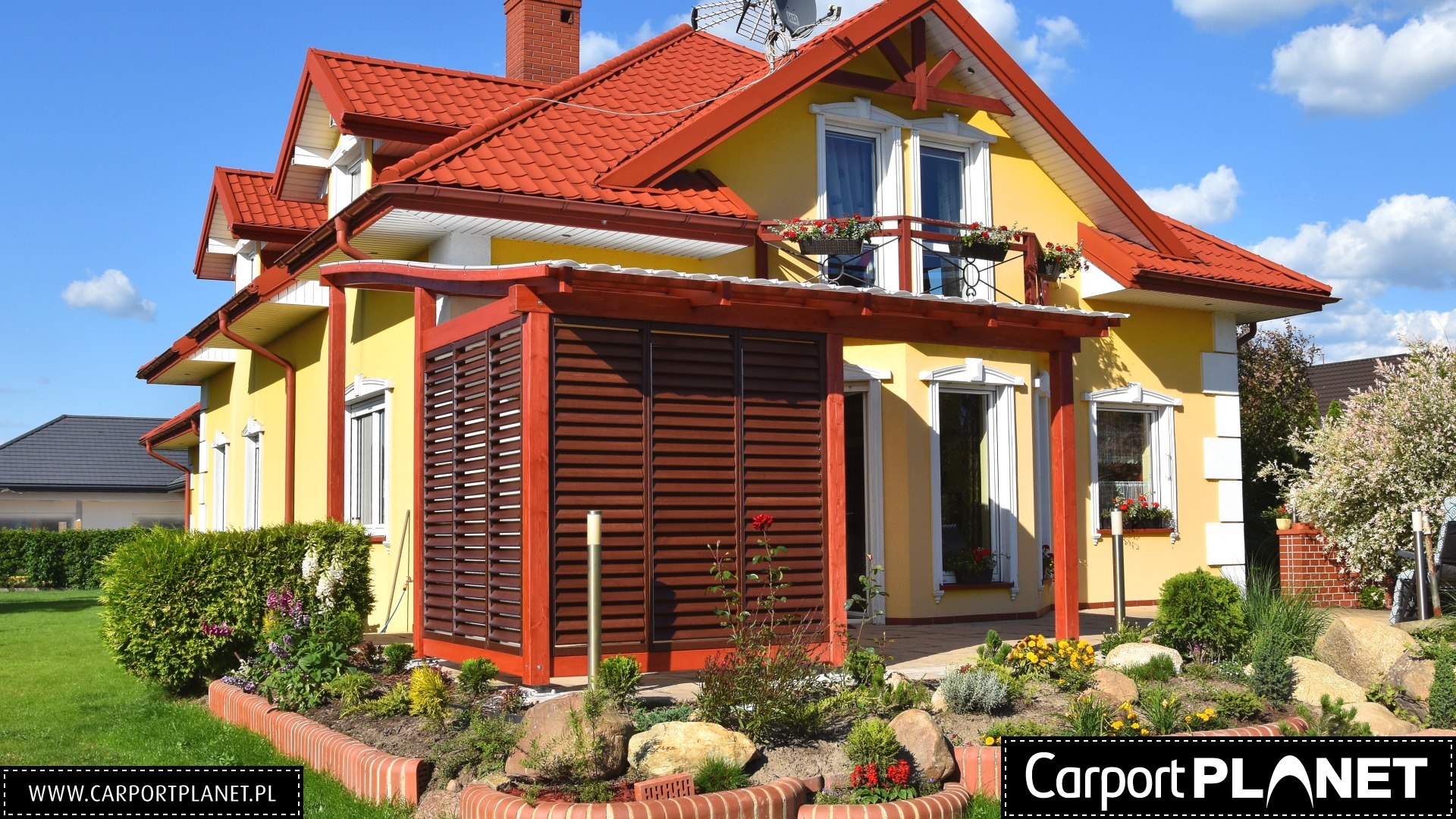 Żółty dom z czerwoną dachówką i drewnianą konstrukcją z żaluzjami, prawdopodobnie osłaniającą taras, otoczony zadbanym ogrodem z kamieniami i ceglanym obrzeżem.
