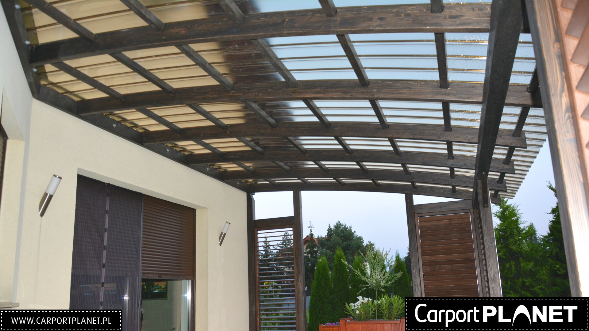 Zadaszenie tarasu z ciemnego drewna z przezroczystym wypełnieniem, widok od dołu, z roletami na ścianie budynku i roślinami w tle.